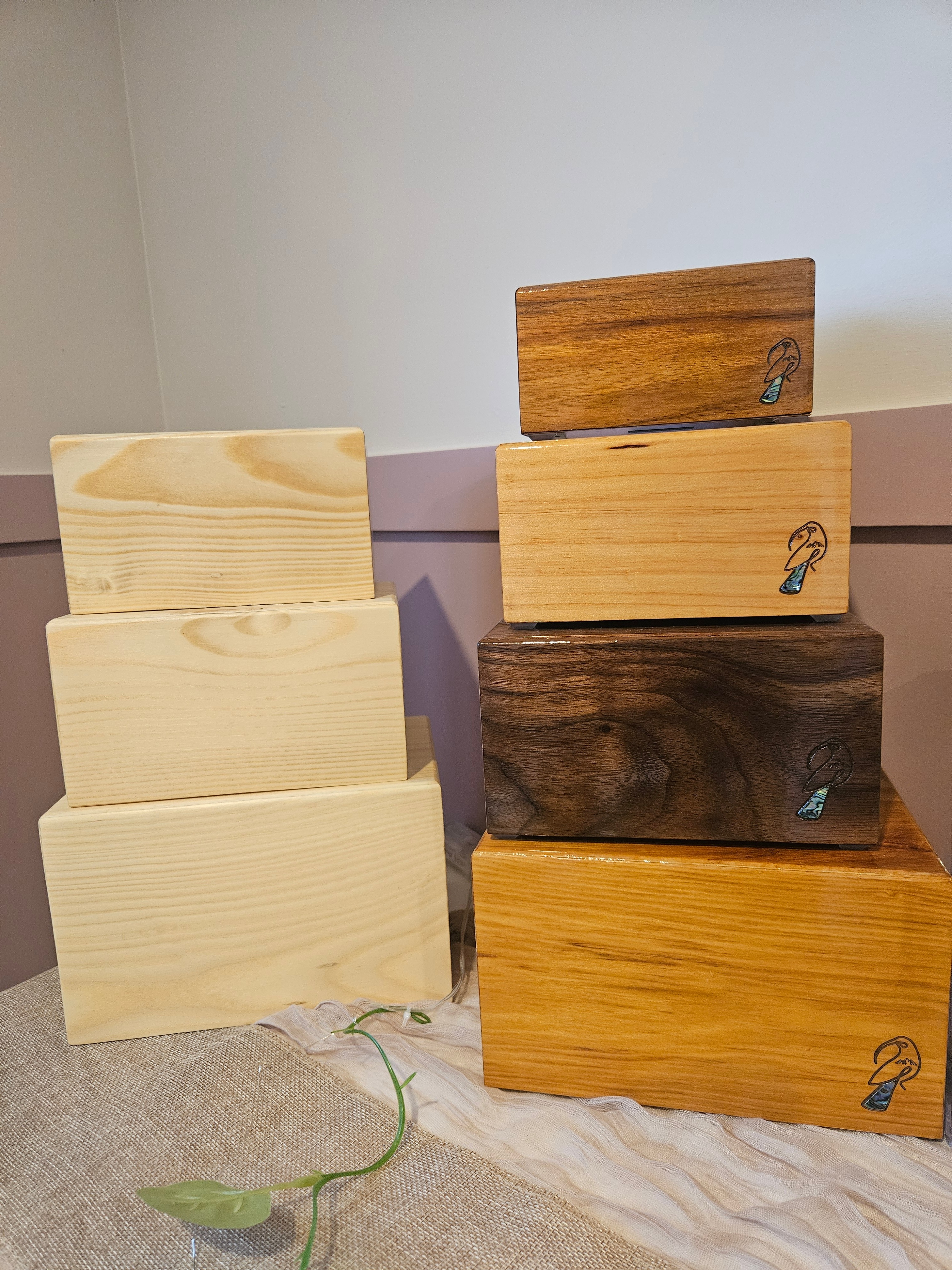 Eco and fibre urns (plain, unfinished pine boxes) on the left, compared with Huia hardwood urns (polished, engraved with a huia bird and paua inlay) on the right